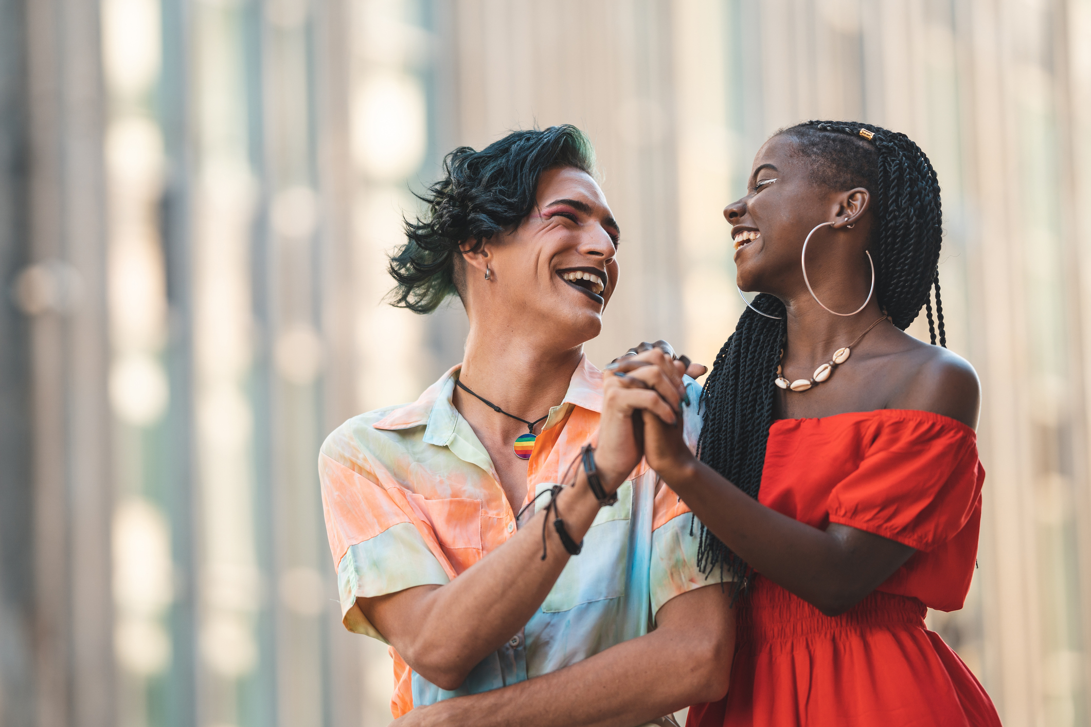 Two trans people, one with pale skin and blue hair and the other with long black braids and darker skin, hold hands and smile happily. 