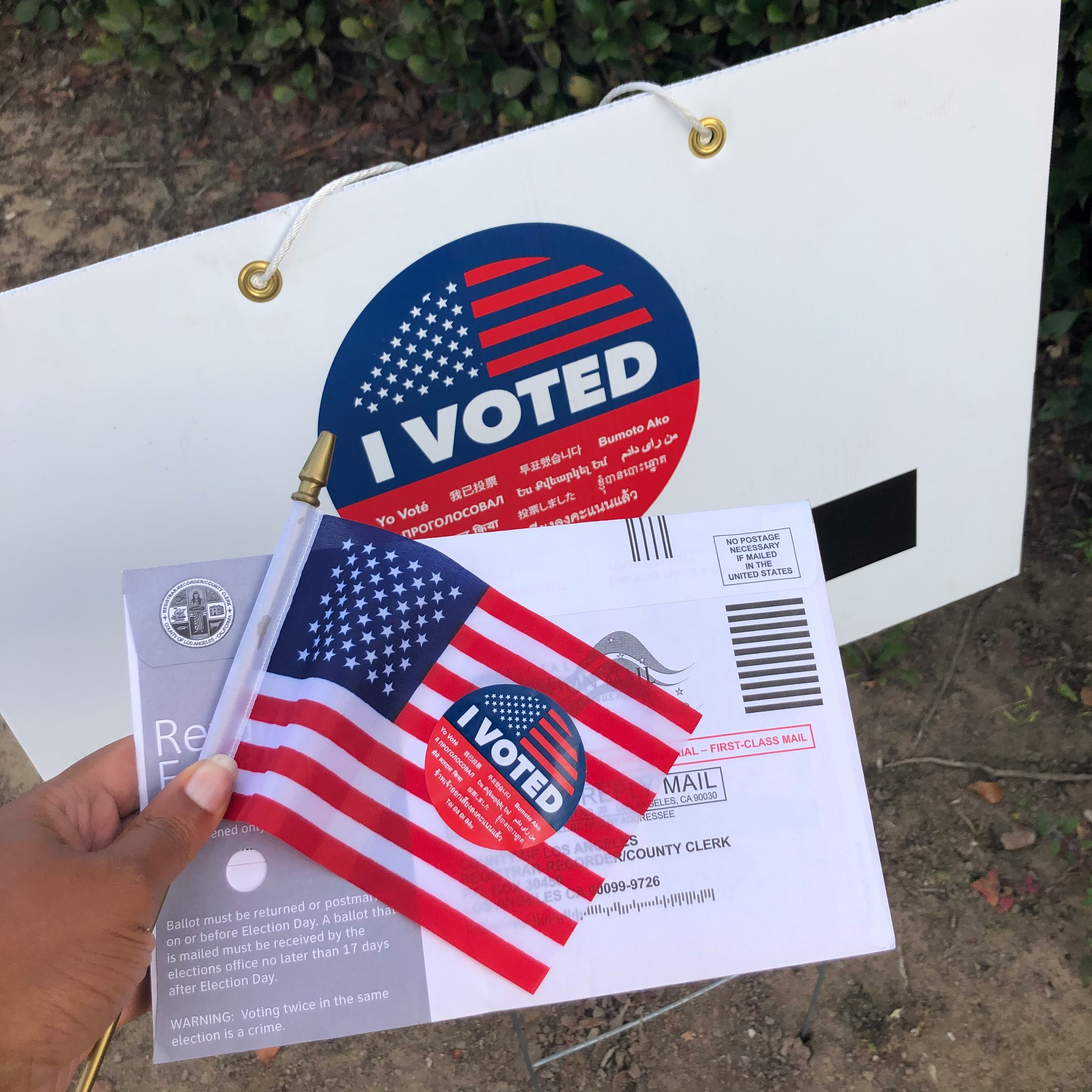 A brown hand holds an American flag and I Voted sticker. 